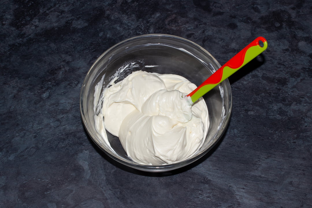Baileys whipped cream being folded into cream cheese in a glass bowl with a green spatula
