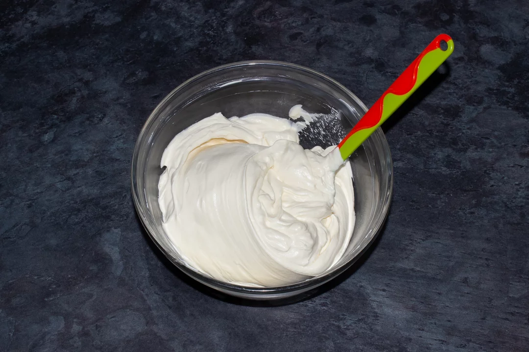 Baileys whipped cream being folded into cream cheese in a glass bowl with a green spatula