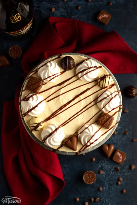 Flat lay view of a Baileys cheesecake on a cake stand set over a deep blue background with a red linen napkin. There are Baileys chocolates and chocolate chips scattered around.