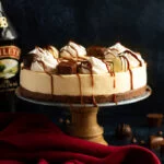Baileys cheesecake on a cake stand set over a deep blue background with a red linen napkin. A bottle of Baileys sits in the background and Baileys chocolates/chocolate chips are scattered around