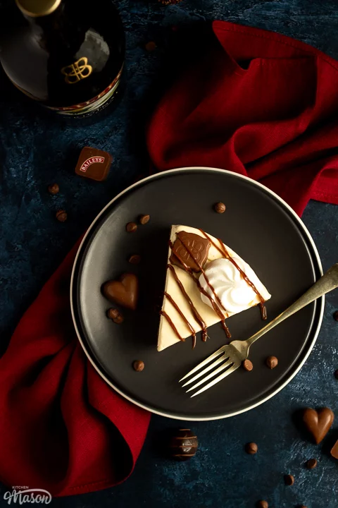 A slice of no bake Baileys cheesecake on a dark grey plate with a fork that's set over a deep blue background and a red linen napkin. There's a bottle of Baileys in the back and Baileys chocolates / chocolate chips scattered around.