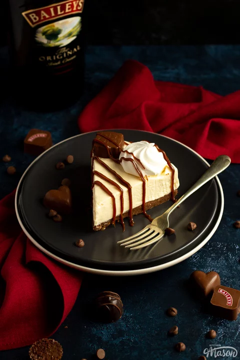 A slice of no bake Baileys cheesecake on a dark grey plate with a fork that's set over a deep blue background and a red linen napkin. There's a bottle of Baileys in the background.