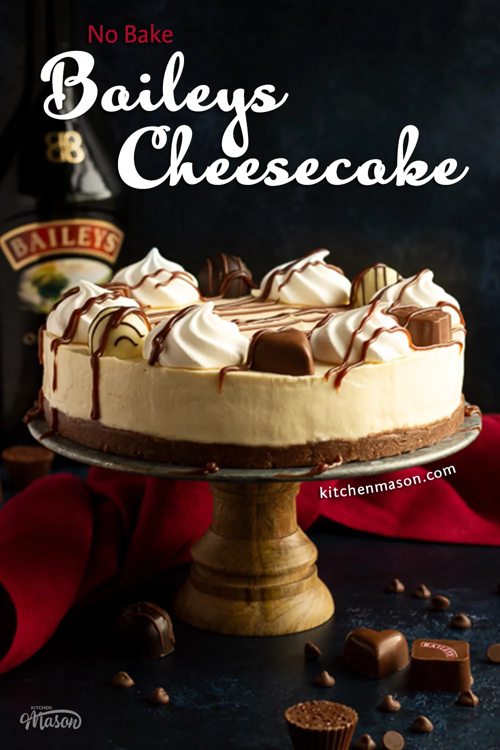 Baileys cheesecake on a cake stand set over a deep blue background with a red linen napkin. A bottle of Baileys sits in the background and Baileys chocolates/chocolate chips are scattered around