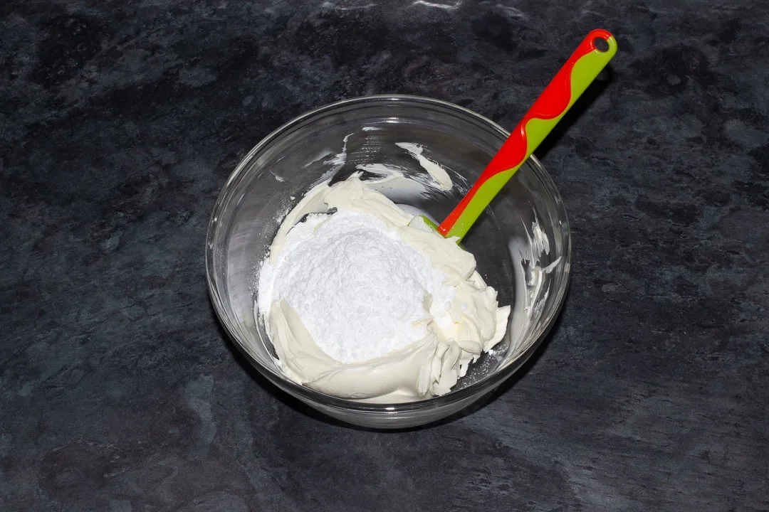 Softened cream cheese and icing sugar in a glass bowl with a green spatula