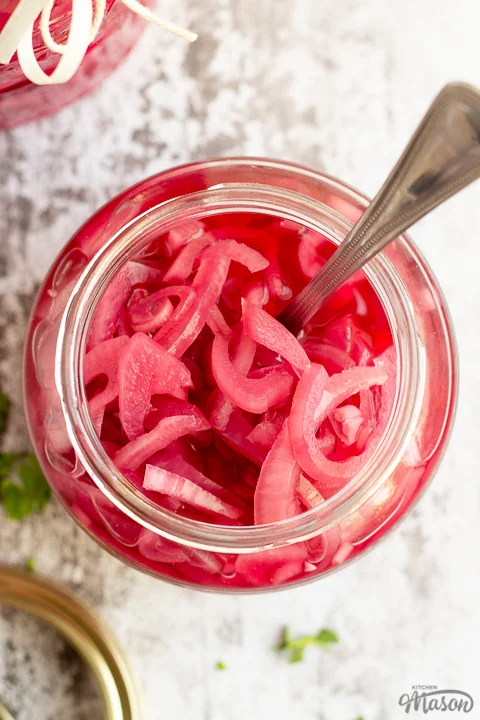 A birds eye view of an open jar of pickled red onions with a fork in it. Set on a rustic white wood backdrop with a sealed jar of pickled red onions, a pickling jar lid and some chopped parsley in the background.