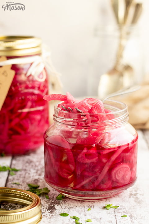 An open jar of pickled red onions with a fork in. Sat on a rustic white wood backdrop with a sealed and labelled jar of pickled red onions, a pickling lid, a jar of cutlery and a light brown napkin in the background.