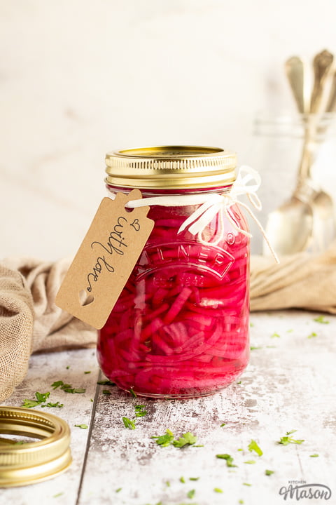 A sealed jar of pickled red onions with a label tied to it that says 'with love'. Sat on a rustic white wood backdrop with a light brown napkin, a pickling jar lid and a jar of cutlery in the background.