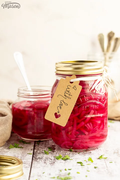 A sealed jar of pickled red onions with a label tied to it that says 'with love'. Sat on a rustic white wood backdrop with an open jar of pickled red onions, a light brown napkin, a pickling jar lid and a jar of cutlery in the background.