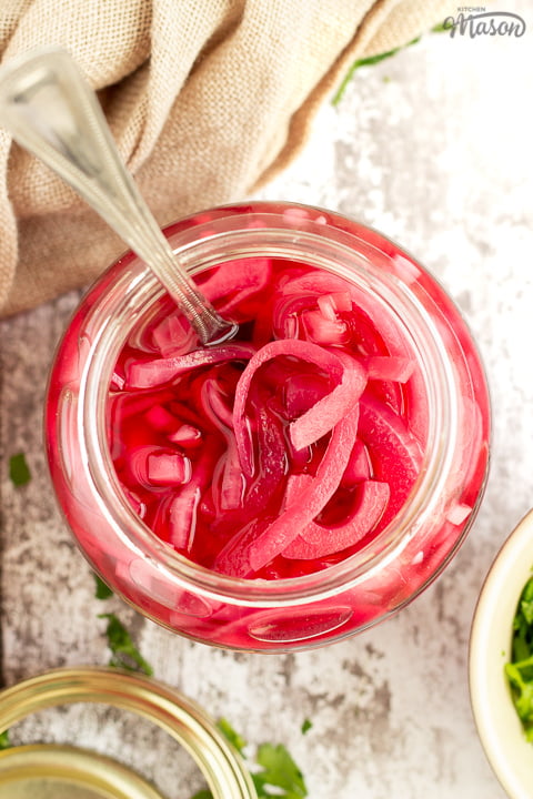 A birds eye view of an open jar of pickled red onions with a fork in it. Set on a rustic white wood backdrop with a light brown napkin, a pickling jar lid and some chopped parsley in the background.