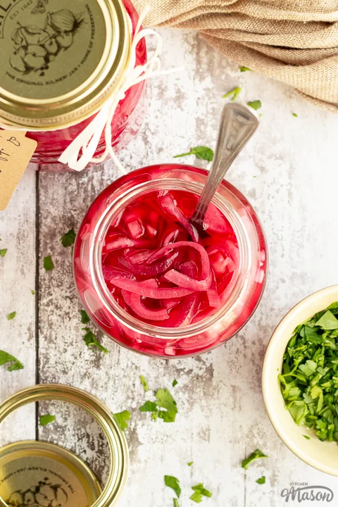 A birds eye view of an open jar of pickled red onions with a fork in it. Set on a rustic white wood backdrop with a sealed jar of pickled red onions, a light brown napkin, a pickling jar lid and some chopped parsley in the background.