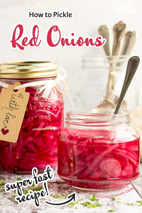 An open jar of pickled red onions with a fork in. Sat on a rustic white wood backdrop with a sealed and labelled jar of pickled red onions, a pickling lid, a jar of cutlery and a light brown napkin in the background.
