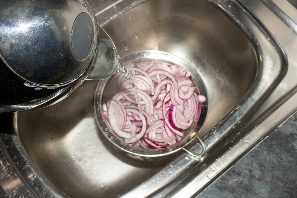Boiling water being poured over sliced red onions in a sieve set over a sink