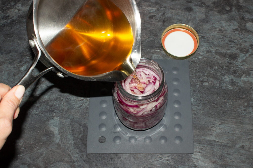 Hot pickling vinegar being poured into a jar of sliced red onions