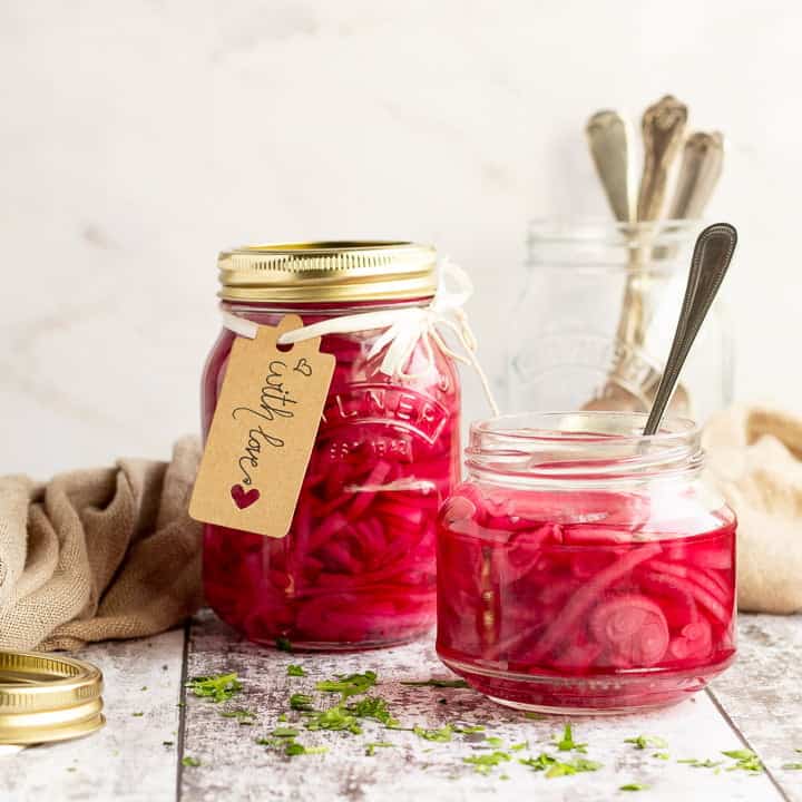 An open jar of pickled red onions with a fork in. Sat on a rustic white wood backdrop with a sealed and labelled jar of pickled red onions, a pickling lid, a jar of cutlery and a light brown napkin in the background.