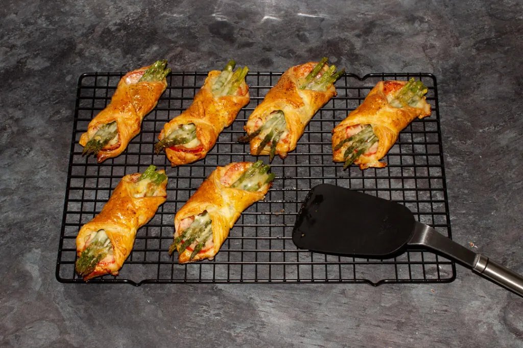 6 baked asparagus puff pastry parcels on a wire rack with a rubber spatula