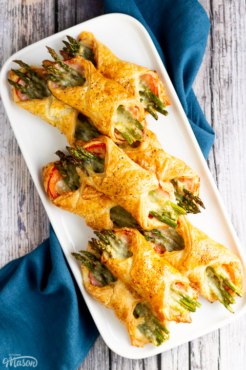 9 asparagus puff pastry parcels on a white rectangular serving plate. Set on a rustic grey wood backdrop with a blue linen napkin.