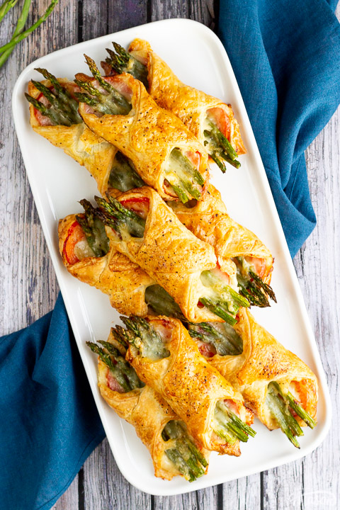 9 asparagus puff pastry parcels on a white rectangular serving plate. Set on a rustic grey wood backdrop with a blue linen napkin and 3 asparagus tips.