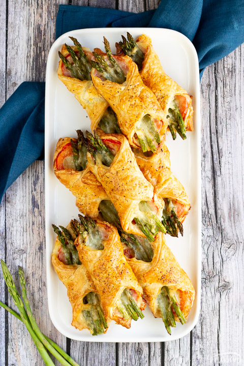 9 asparagus puff pastry parcels on a white rectangular serving plate. Set on a rustic grey wood backdrop with a blue linen napkin and 3 asparagus tips.
