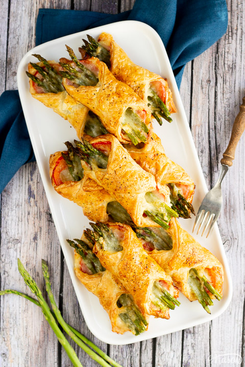 9 asparagus puff pastry parcels on a white rectangular serving plate. Set on a rustic grey wood backdrop with a blue linen napkin, 3 asparagus tips and a wood handled fork.