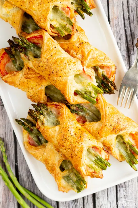 9 asparagus puff pastry parcels on a white rectangular serving plate. Set on a rustic grey wood backdrop with 3 asparagus tips and a wood handled fork.