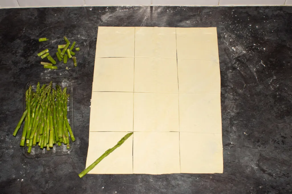 A sheet of ready rolled puff pastry cur into squares on a lightly floured work surface. There are asparagus tips being cut to size too.