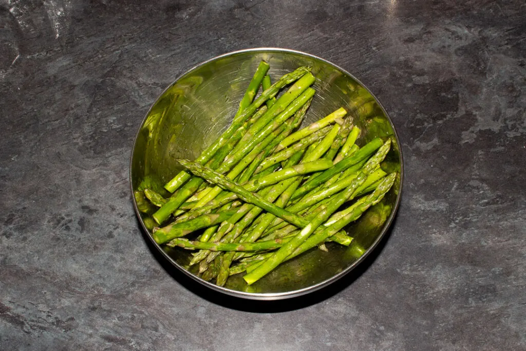 Asparagus tips coated in olive oil, salt and pepper in a metal bowl on a work surface