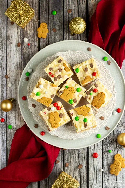 Christmas rocky road squares on a lace white plate set on a red linen napkin over a grey wood backdrop. There are Christmas sprinkles, gold baubles and decorations scattered around.