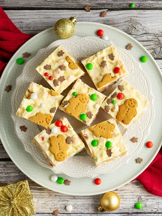 Christmas rocky road squares on a lace white plate set on a red linen napkin over a grey wood backdrop. There are Christmas sprinkles, gold baubles and decorations scattered around.