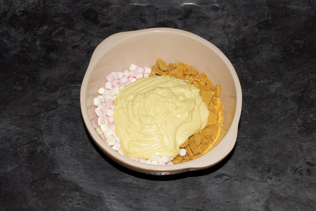 Pink and white mini marshmallows and crushed gingerbread biscuits in a large bowl with melted white chocolate poured on top on a kitchen worktop