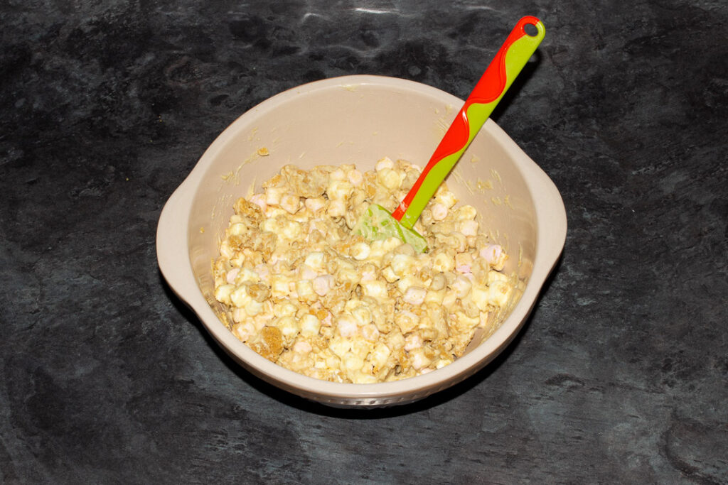 Mini marshmallows, crushed ginger biscuits and melted white chocolate mixed together in a large bowl on a kitchen worktop with a green spatula