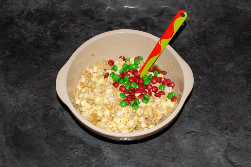 Mini marshmallows, crushed ginger biscuits and melted white chocolate mixed together in a large bowl with green and red M&M's on top on a kitchen worktop with a green spatula