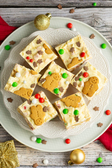 Christmas rocky road squares on a lace white plate set on a red linen napkin over a grey wood backdrop. There are Christmas sprinkles, gold baubles and decorations scattered around.