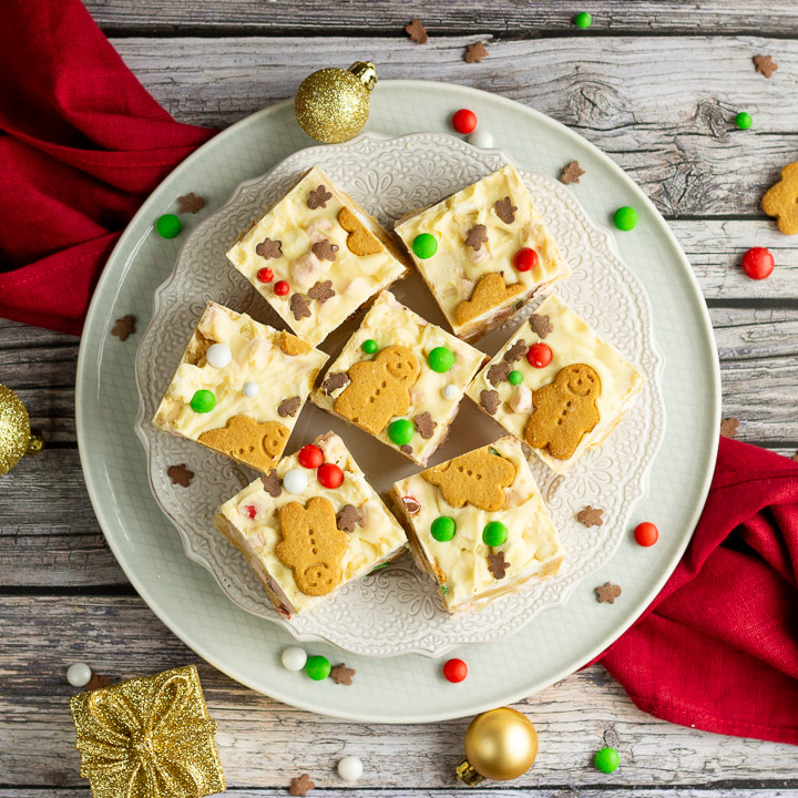 Christmas rocky road squares on a lace white plate set on a red linen napkin over a grey wood backdrop. There are Christmas sprinkles, gold baubles and decorations scattered around.