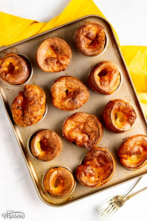 Birds eye view of Yorkshire puddings in a Yorkshire pudding pan with a mustard yellow napkin and 2 forks in the background.