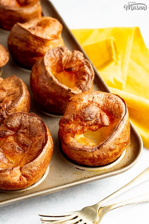 Yorkshire puddings in a Yorkshire pudding pan with a mustard yellow napkin and 2 forks in the background.