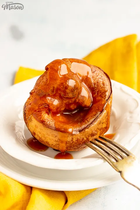 A Yorkshire pudding with gravy poured over it in a small white bowl set over a mustard yellow napkin. There's a fork resting on the bowl.