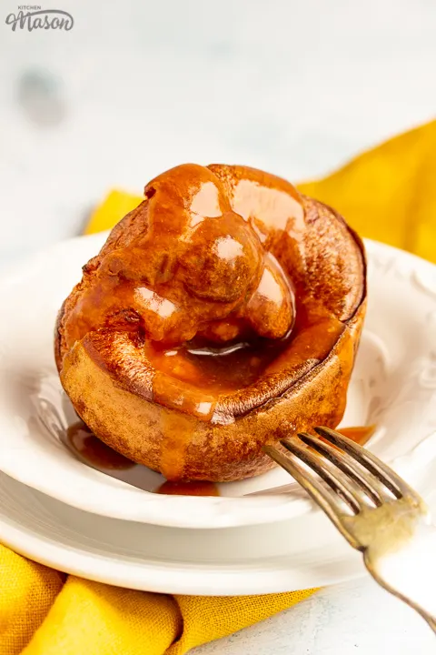 A Yorkshire pudding with gravy poured over it in a small white bowl set over a mustard yellow napkin. There's a fork resting on the bowl.