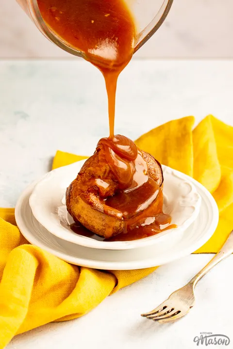 A Yorkshire pudding with gravy being poured over it in a small white bowl set over a mustard yellow napkin with a fork by it's side.
