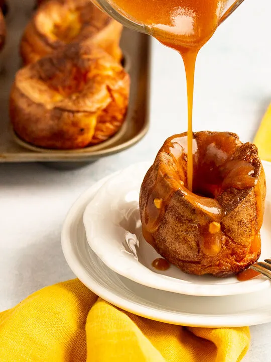 A Yorkshire pudding with gravy being poured over it in a small white bowl set over a mustard yellow napkin. There's a fork resting on the bowl and more Yorkshire puddings in a Yorkshire pudding tin in the background.