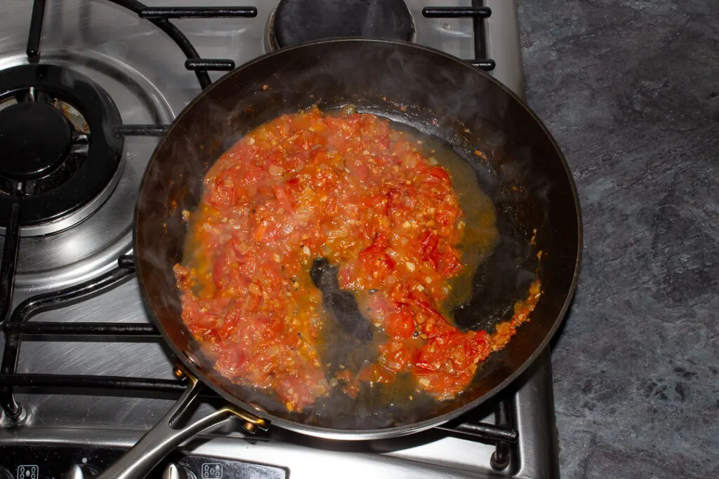Cooked onion, garlic, cherry tomatoes and balsamic vinegar in a frying pan on a stove with pasta water added.