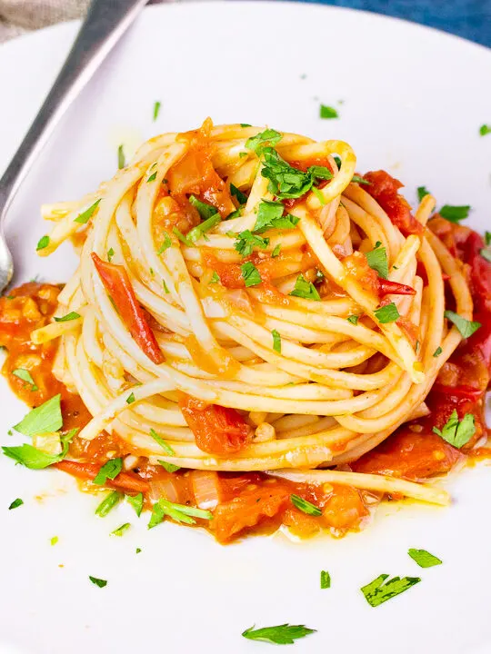 A close up side view of a nest of fresh tomato spaghetti on a white plate scattered with chopped parsley. There's a fork resting on the plate and a light brown napkin in the background, all set on a blue backdrop.