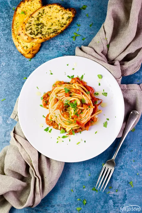 A birds eye view of a nest of fresh tomato spaghetti on a white plate scattered with chopped parsley. There's some garlic bread slices, a fork and a light brown napkin in the background, all set on a blue backdrop.