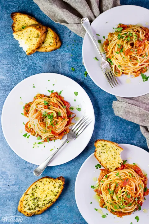 A birds eye view of 3 plates with nests of fresh tomato spaghetti on white plates scattered with chopped parsley. There's some garlic bread slices, forks and a light brown napkin in the background, all set on a blue backdrop.