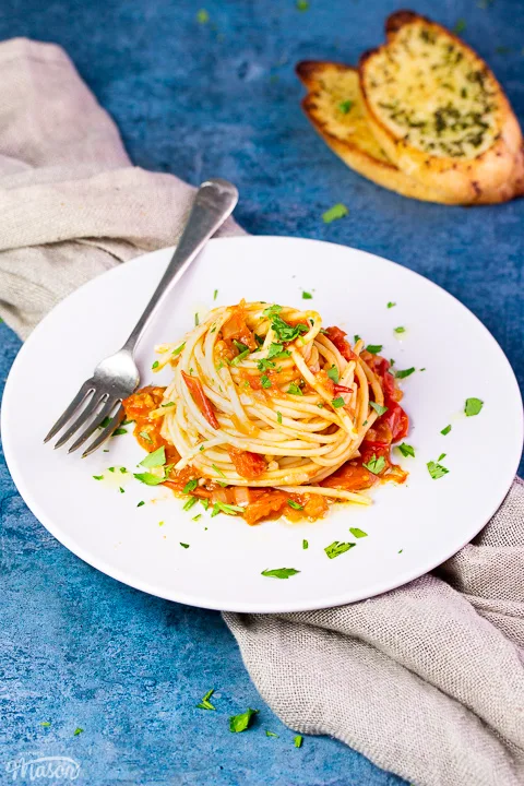 A side view of a nest of fresh tomato spaghetti on a white plate scattered with chopped parsley. There's some garlic bread slices, a fork and a light brown napkin in the background, all set on a blue backdrop.