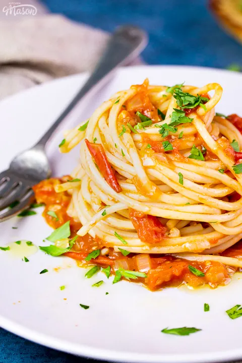 A close up side view of a nest of tomato spaghetti on a white plate scattered with chopped parsley. There's a fork resting on the plate and it's set on a blue backdrop.
