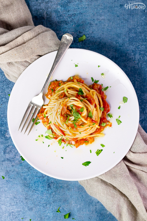 A birds eye view of a nest of fresh tomato spaghetti on a white plate scattered with chopped parsley. There's a fork resting on the plate and a light brown napkin in the background, all set on a blue backdrop.