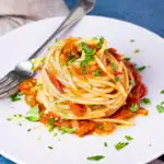 A side view of a nest of fresh tomato spaghetti on a white plate scattered with chopped parsley. There's a fork resting on the plate and a light brown napkin in the background, all set on a blue backdrop.