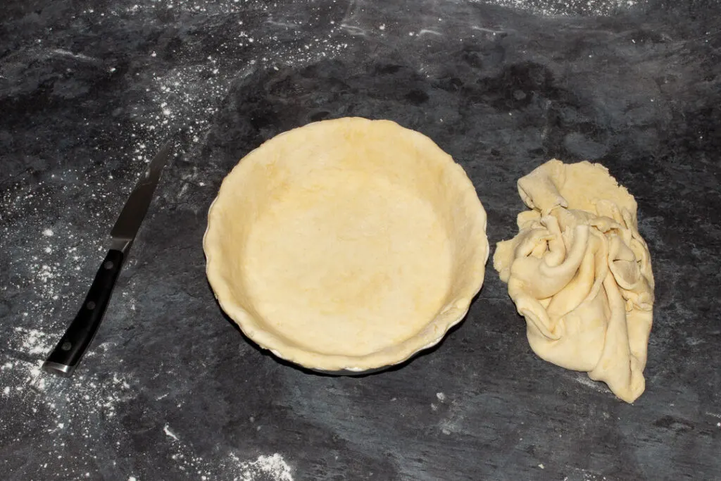A pie dish lined with shortcrust pastry, trimmed excess pastry on the worktop next to it