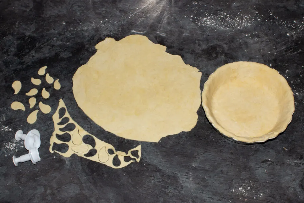 A pie dish lined with shortcrust pastry, remaining pastry rolled out ready to cut the pie lid and some pastry scraps having leaves cut out of them, all on a floured worktop
