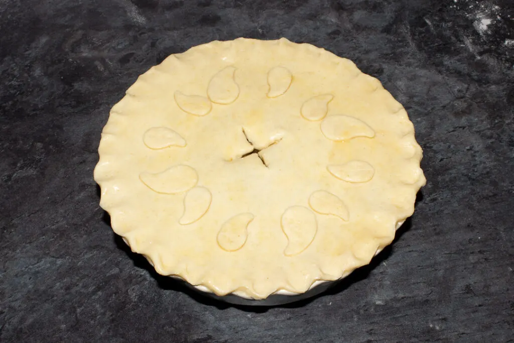 An unbaked vegetarian mince and onion pie with crimped edges, decorated with pastry leaves and brushed all over with egg